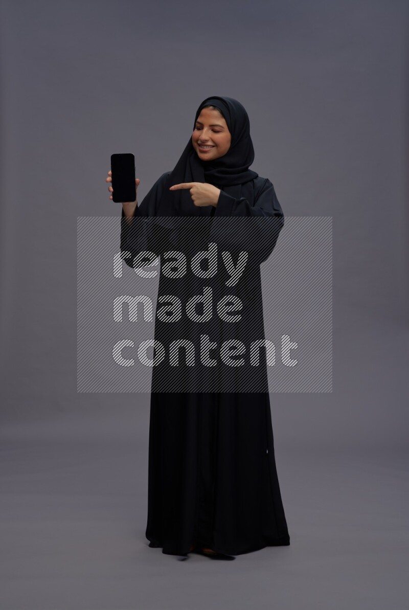 Saudi woman wearing Abaya standing showing phone to camera on gray background