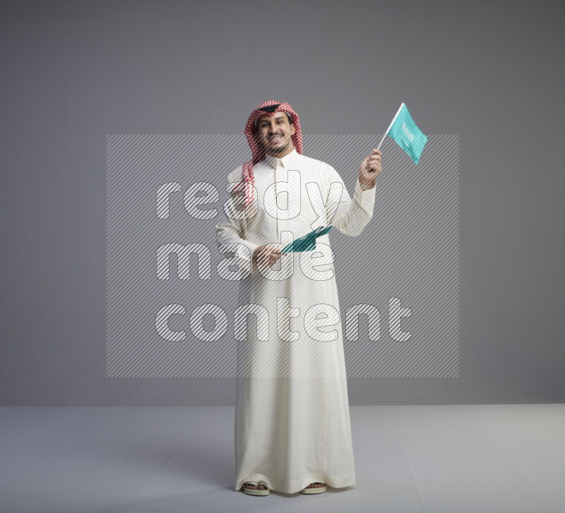 A Saudi man standing wearing thob and red shomag raising small Saudi flag on gray background