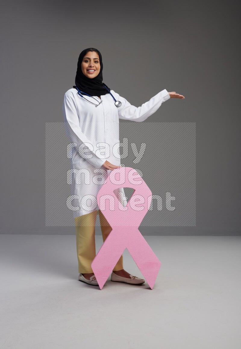 Saudi woman wearing lab coat with stethoscope standing holding awareness ribbon symbol standing on Gray background
