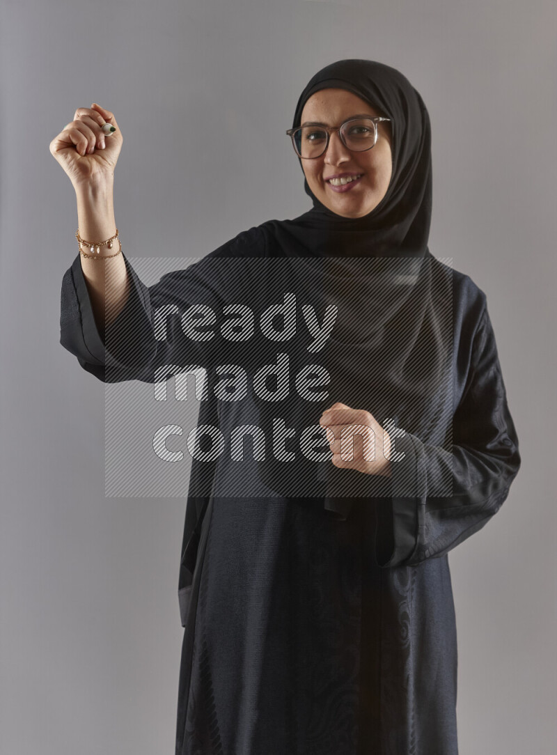 Woman in black abaya, hijab and glasses holding a marker pen to write on a transparent board with different reactions and expressions on grey background