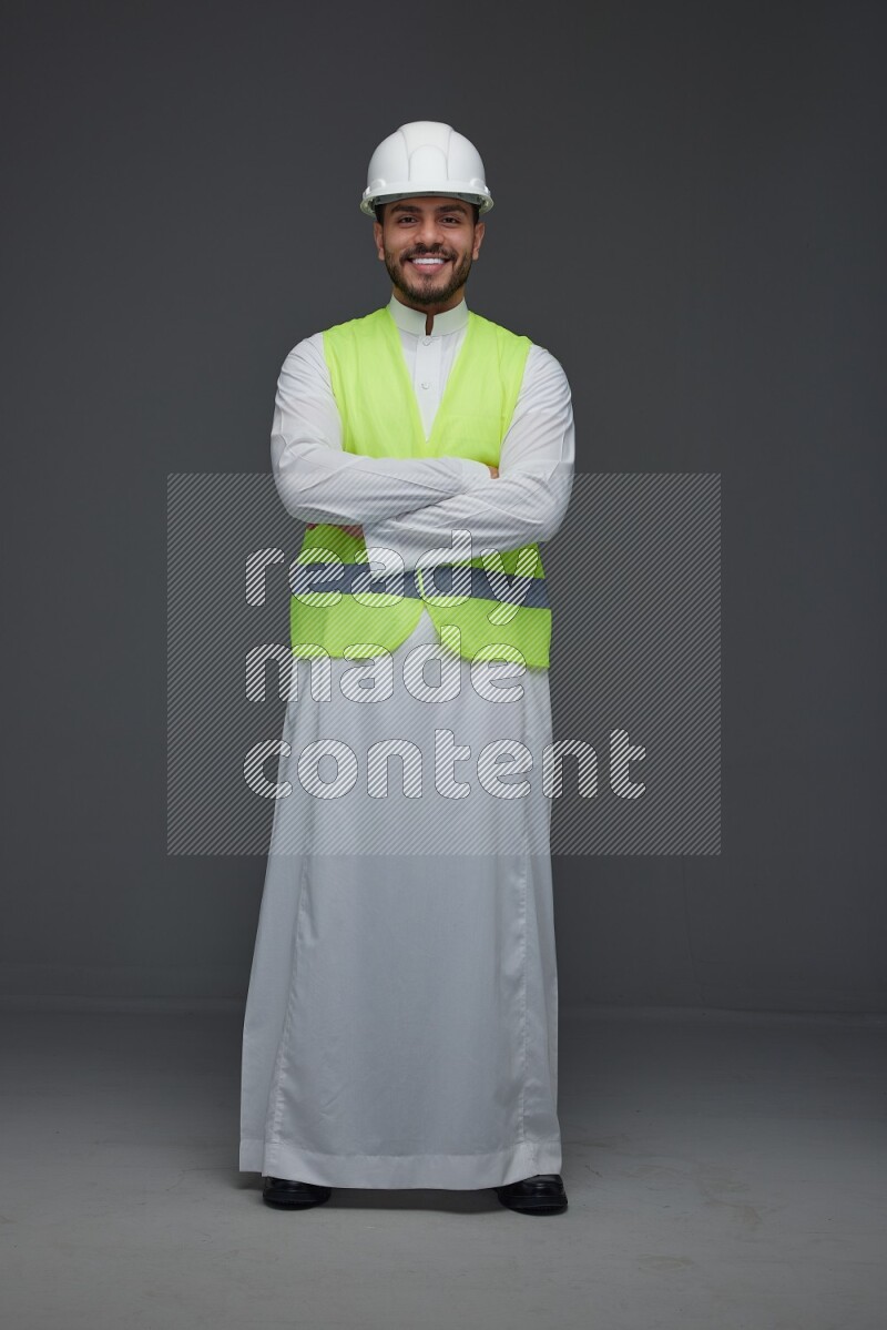 A Saudi man wearing Thobe with a yellow safety vest and white helmet standing and crossing his hands eye level on a gray background
