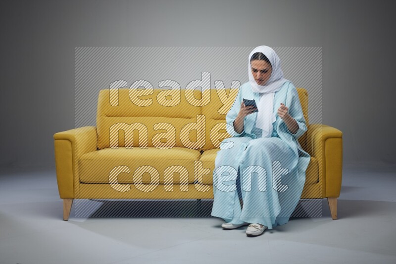 A Saudi woman wearing a light blue Abaya and white head scarf sitting on a yellow sofa and using her phone eye level on a grey background