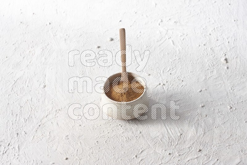 Ceramic beige bowl full of cinnamon powder with a wooden spoon on a textured white background