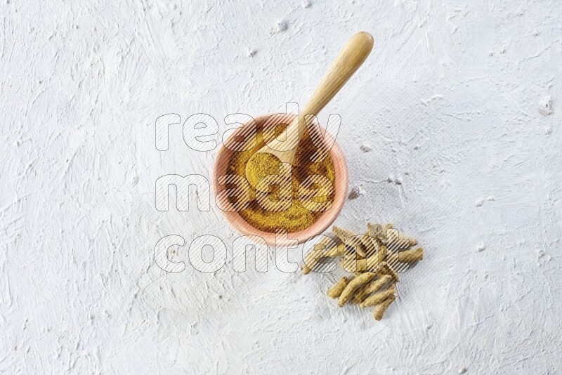 A wooden bowl and wooden spoon full of turmeric powder with dried turmeric fingers beside it on textured white flooring