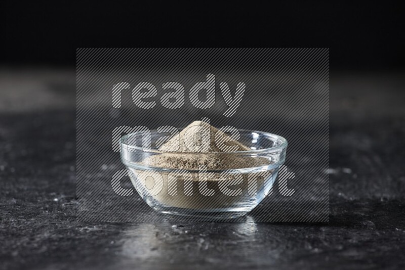 A glass bowl full of white pepper powder on textured black flooring