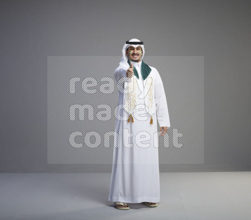 A Saudi man standing wearing thob and white shomag with face painting and Saudi flag scarf on gray background