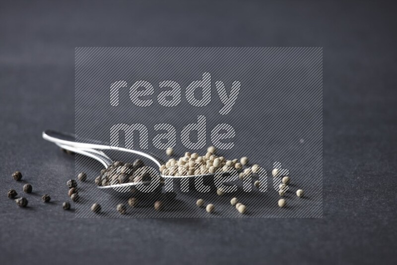 ملعقتان معدنيتان ممتلئتان بحبوب الفلفل الأسود والأبيض مع حبوب متناثرة على أرضية سوداء