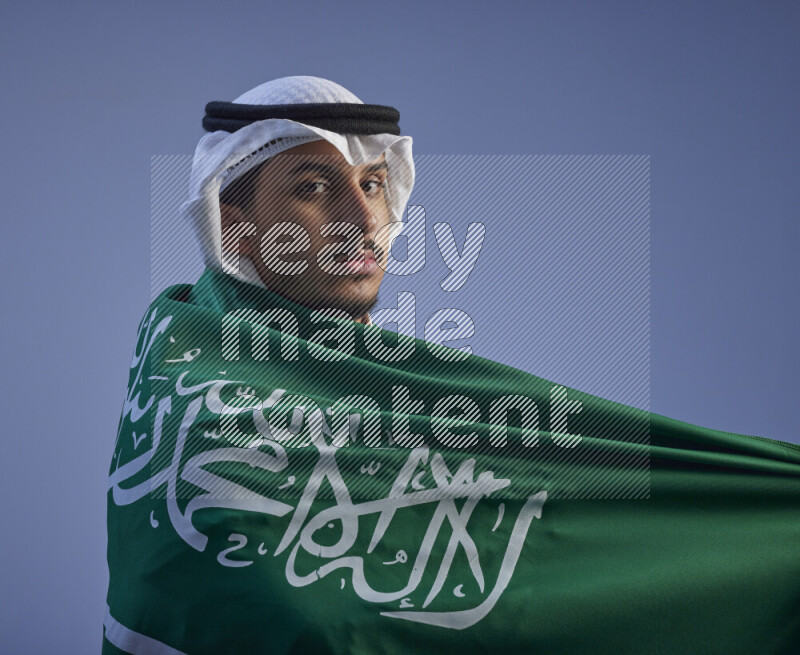 A close-up shot of Saudi man wearing thob and white shomag wrapping big Saudi flag on gray background
