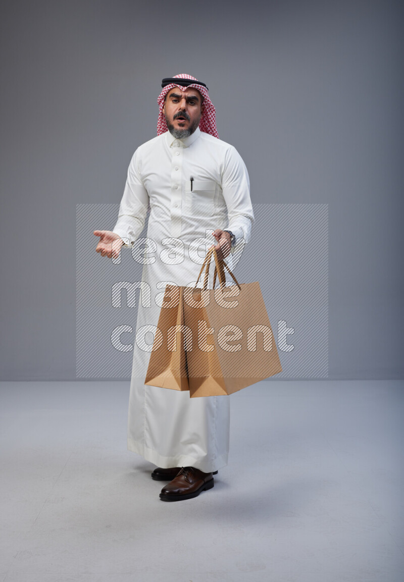 Saudi man Wearing Thob and red Shomag standing holding shopping bag on Gray background