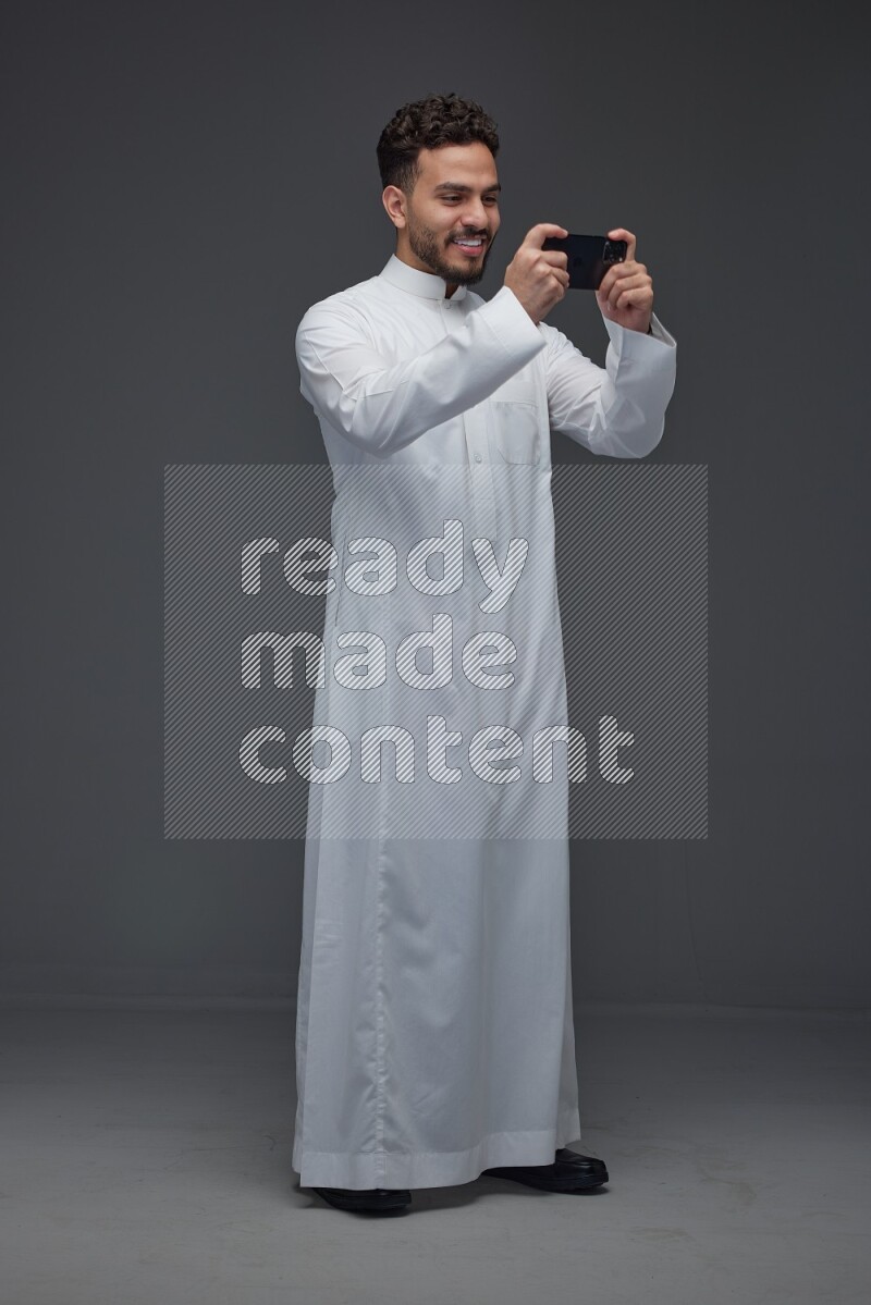 A Saudi man wearing Thobe and playing using his phone horizontally playing video games while standing and making different poses eye level on a gray background