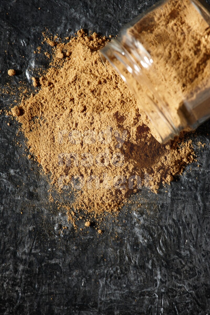 A flipped glass jar full of allspice powder and powder spilled out of it on a textured black flooring
