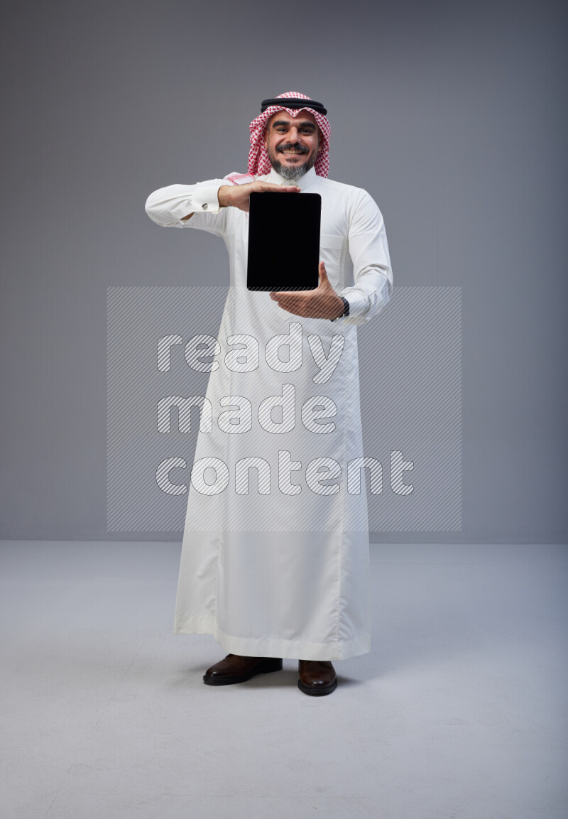 Saudi man Wearing Thob and red Shomag standing showing tablet to camera on Gray background