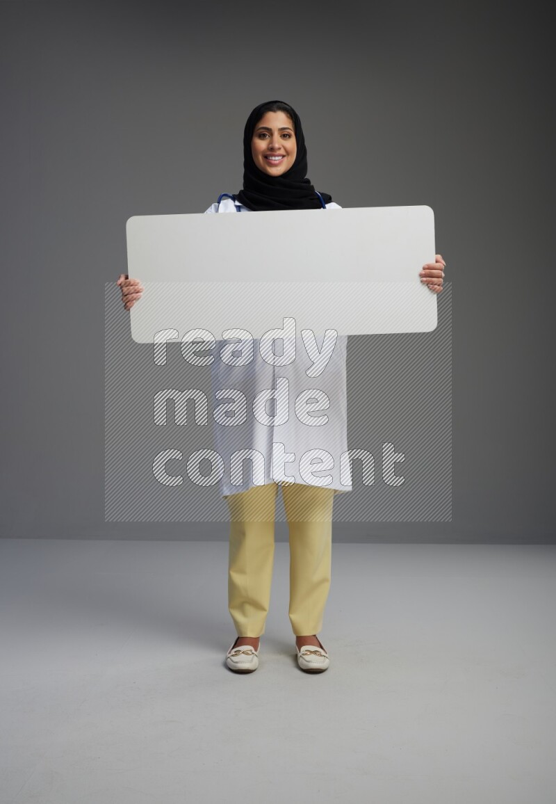 Saudi woman wearing lab coat with stethoscope standing holding board on Gray background