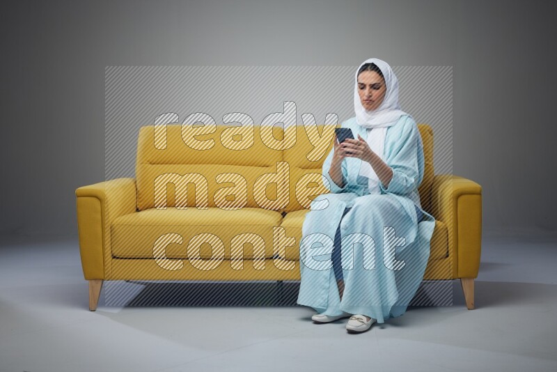A Saudi woman wearing a light blue Abaya and white head scarf sitting on a yellow sofa and texting on her phone on a grey background