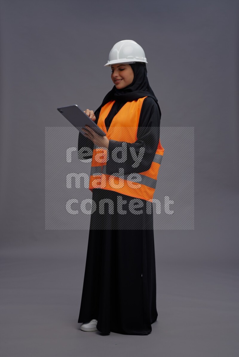 Saudi woman wearing Abaya with engineer vest standing working on tablet on gray background