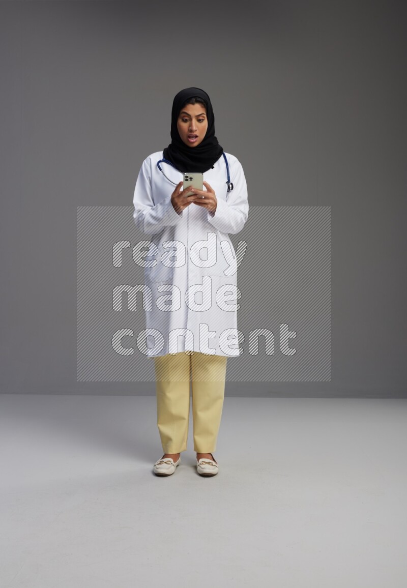 Saudi woman wearing lab coat with stethoscope standing texting on phone on Gray background