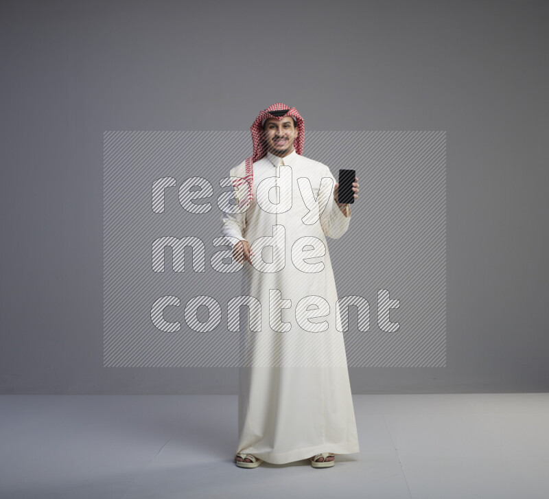 A Saudi man standing wearing thob and red shomag showing phone to camera on gray background