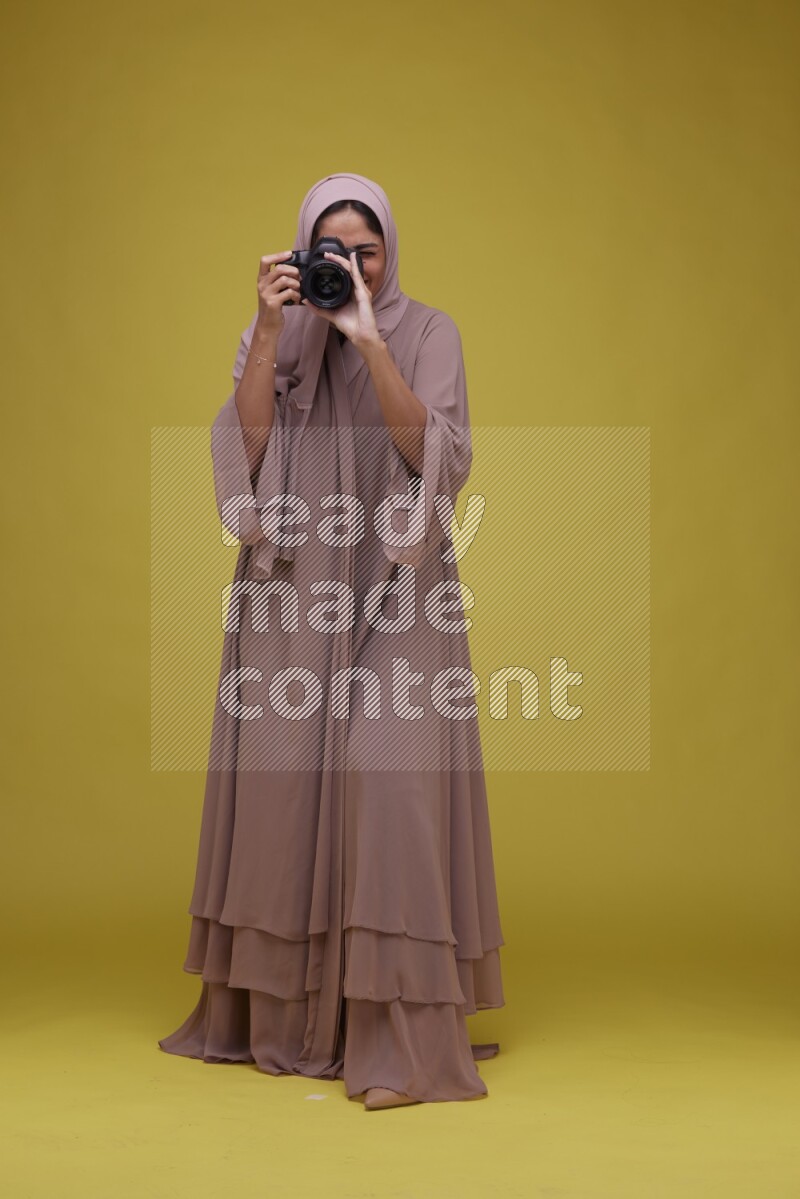 A woman Holding a Camera on a Yellow Background wearing Brown Abaya with Hijab