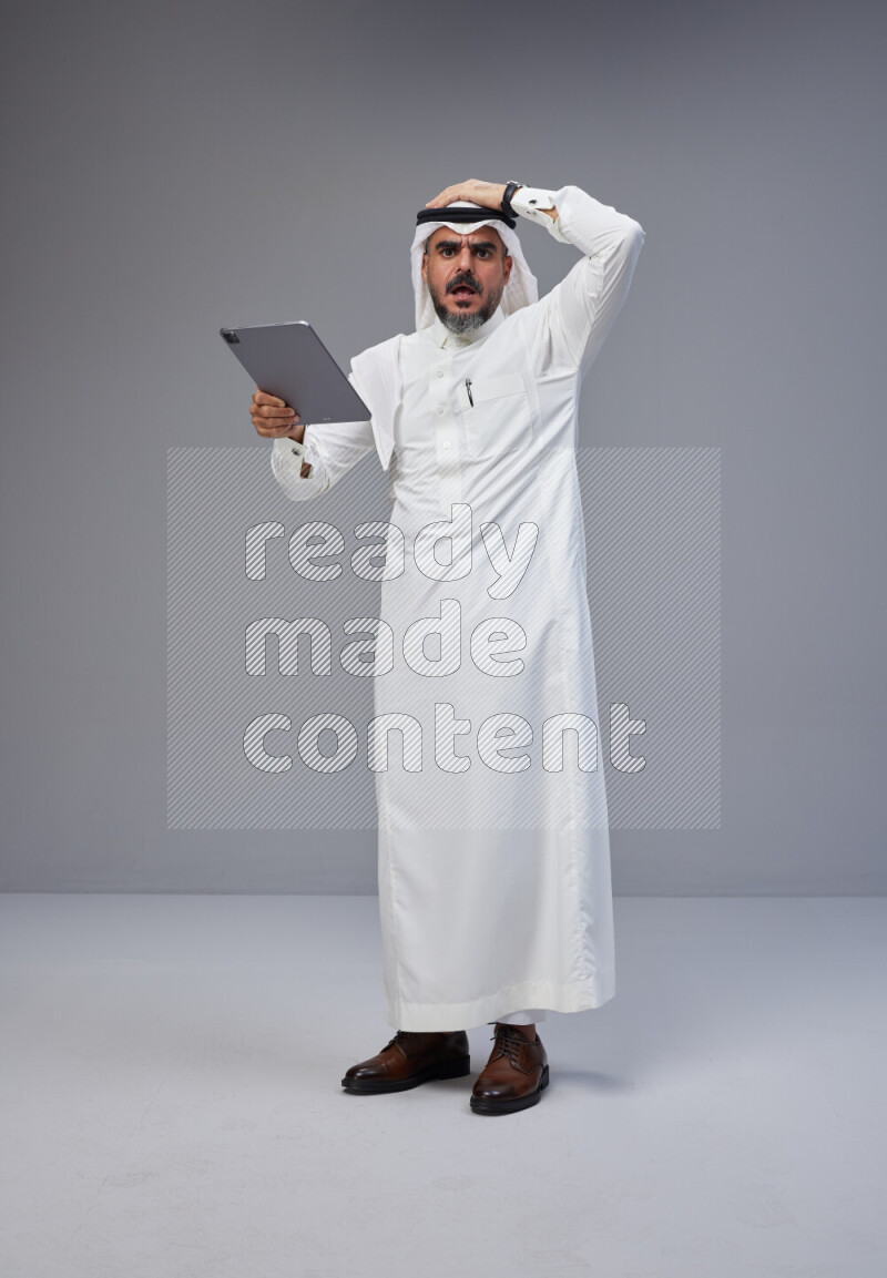 Saudi man Wearing Thob and white Shomag standing working on tablet on Gray background