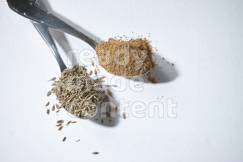 2 metal spoons full of cumin seeds and powder on white flooring