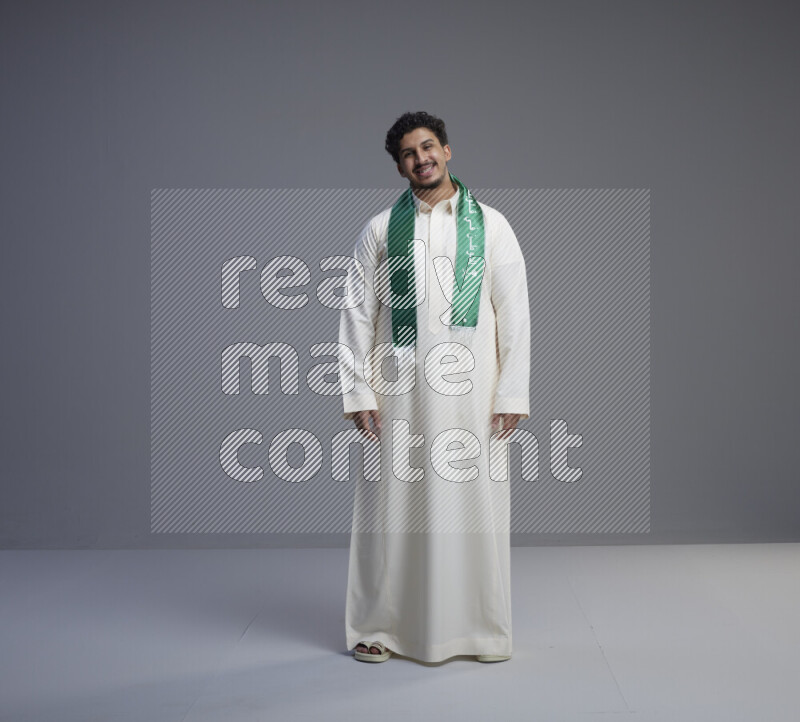 A Saudi man standing wearing thob and Saudi flag scarf on gray background