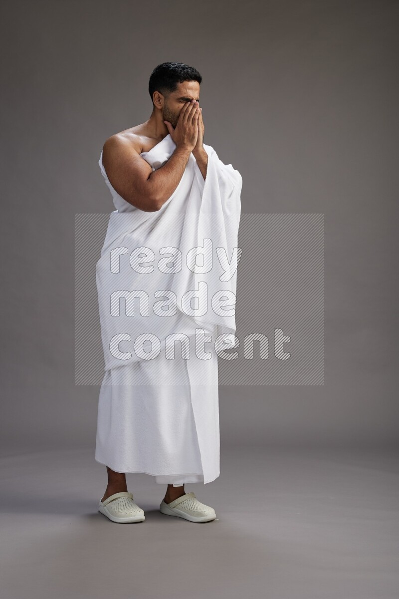 A man wearing Ehram standing showing how to sneeze on gray background