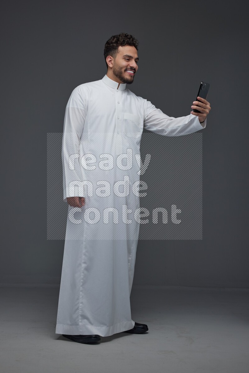A Saudi man wearing Thobe and taking selfie with his phone making different poses eye level on a gray background