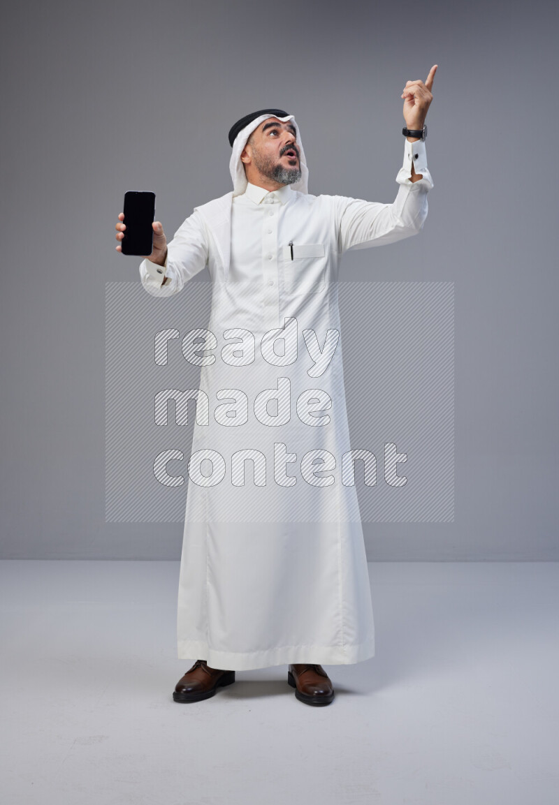 Saudi man Wearing Thob and white Shomag standing showing phone to camera on Gray background