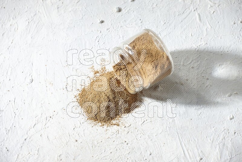 A flipped glass jar full of cumin powder and powder spilled out on textured white flooring