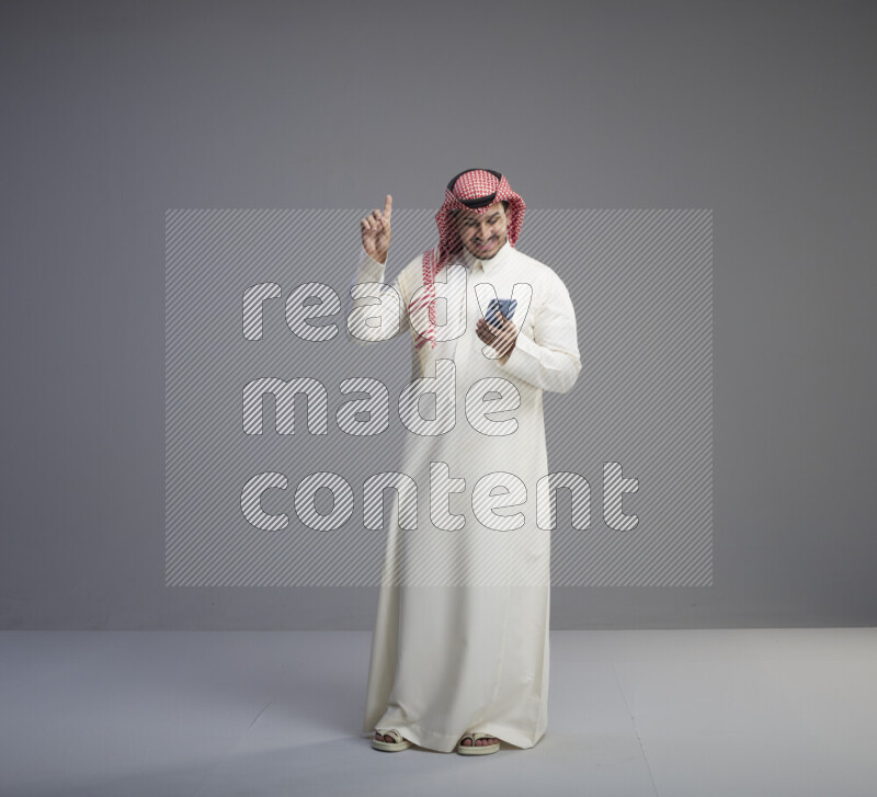 A  Saudi man standing wearing thob and red shomag texting on phone on gray background