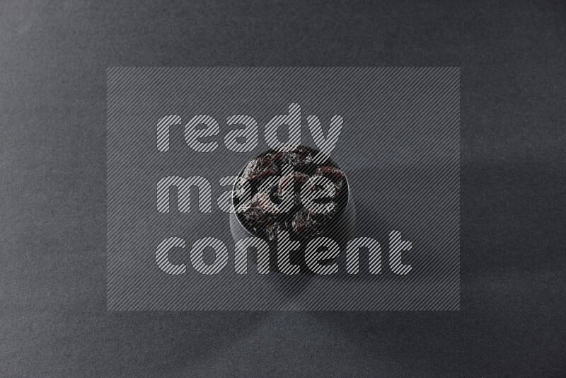 A black pottery bowl full of dried plums on a black background in different angles