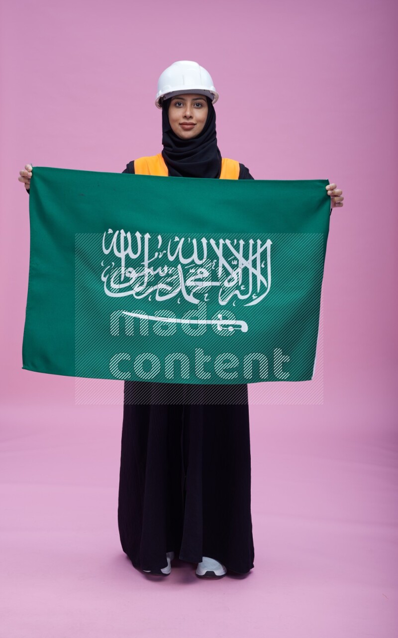 Saudi woman wearing Abaya with engineer vest and helmet standing holding Saudi flag on pink background