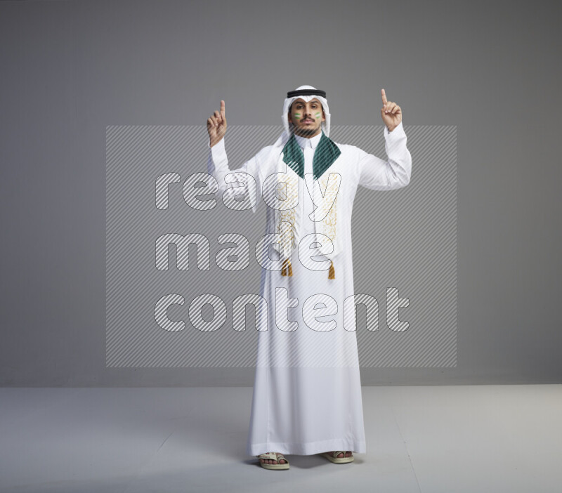 A Saudi man standing wearing thob and white shomag with face painting and Saudi flag scarf on gray background