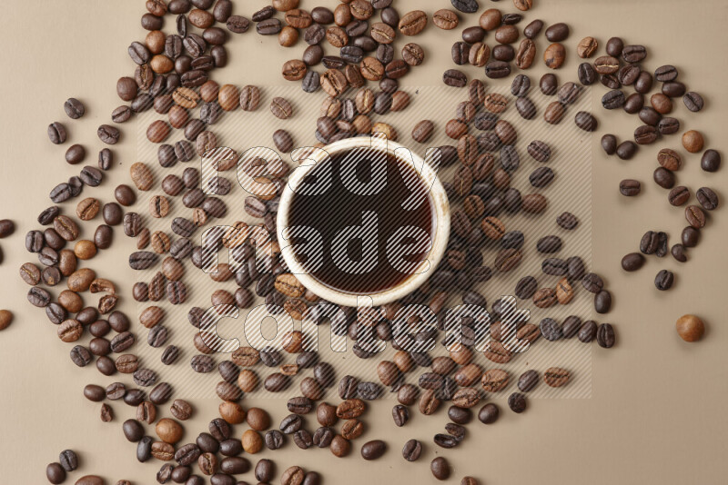 A beige pottery cup of coffee surrounded by roasted coffee beans on beige background