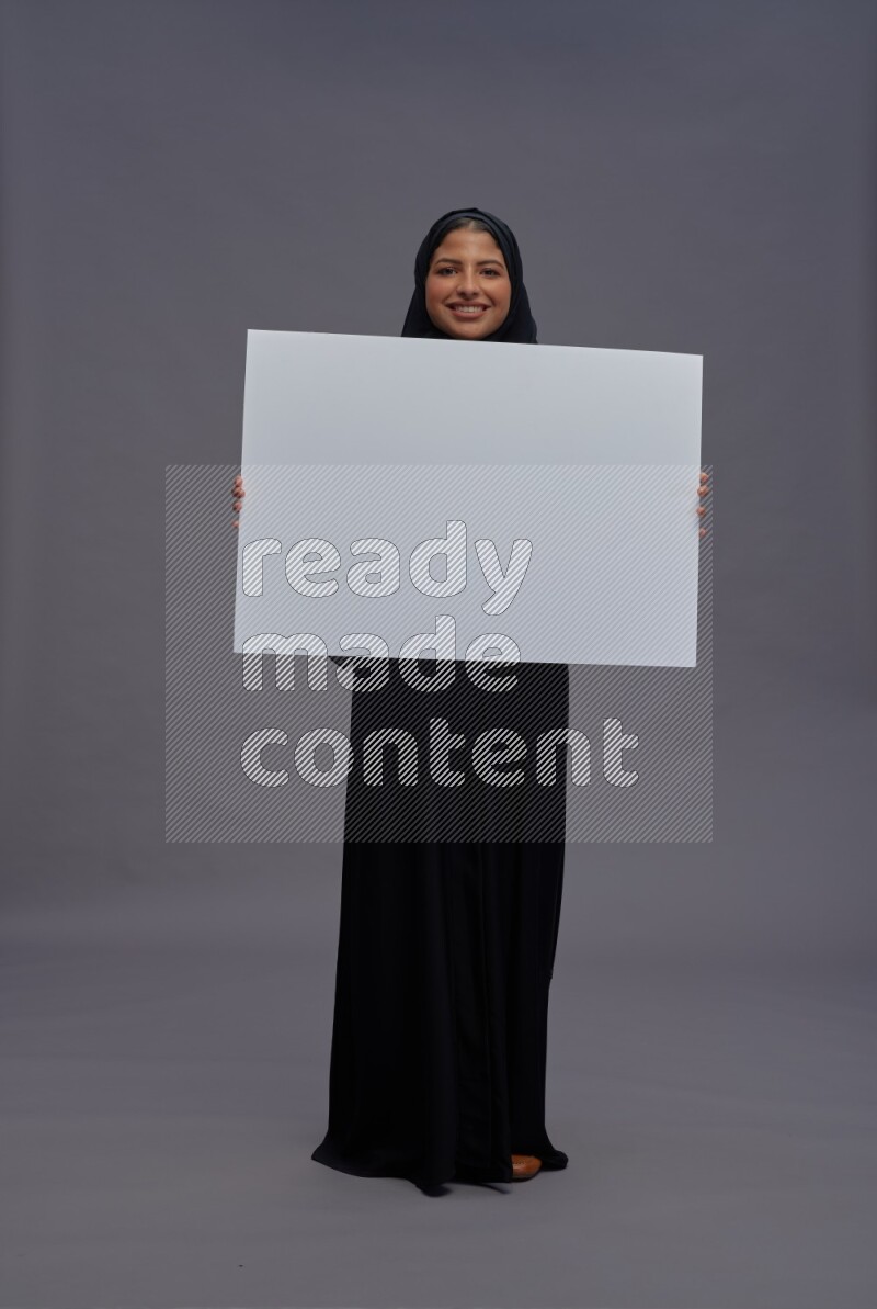Saudi woman wearing Abaya standing holding white board on gray background