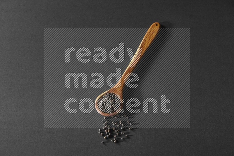 A wooden ladle full of black pepper beads on a black flooring