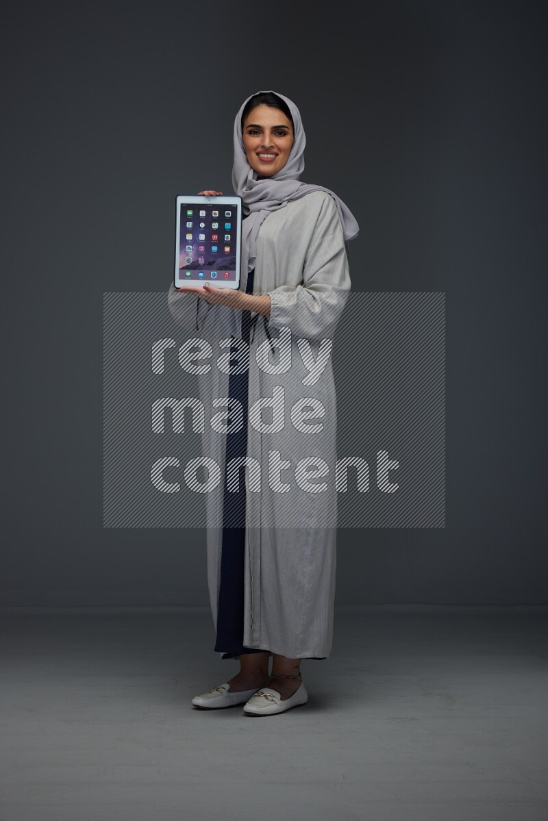 A Saudi woman wearing a light gray Abaya and head scarf standing and showing the phone's screen eye level on a grey background