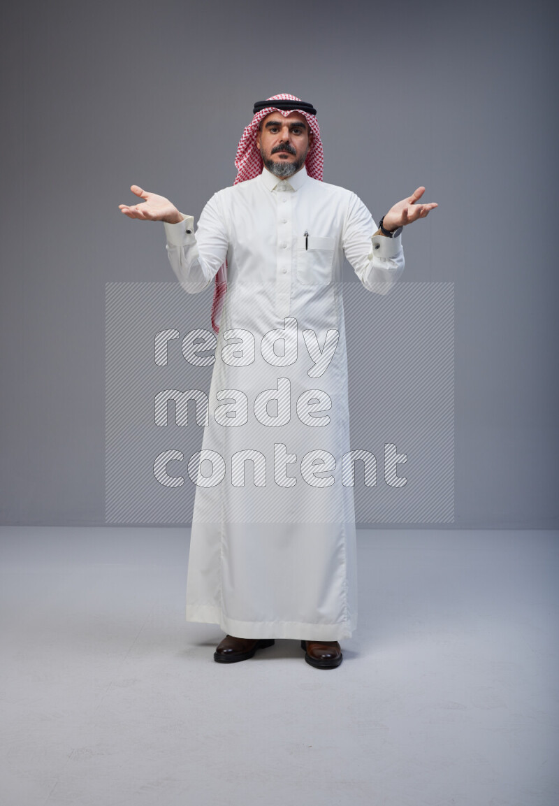 Saudi man Wearing Thob and red white Shomag standing interacting with the camera on Gray background