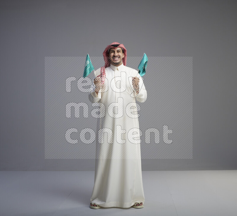 A Saudi man standing wearing thob and red shomag raising small Saudi flag on gray background