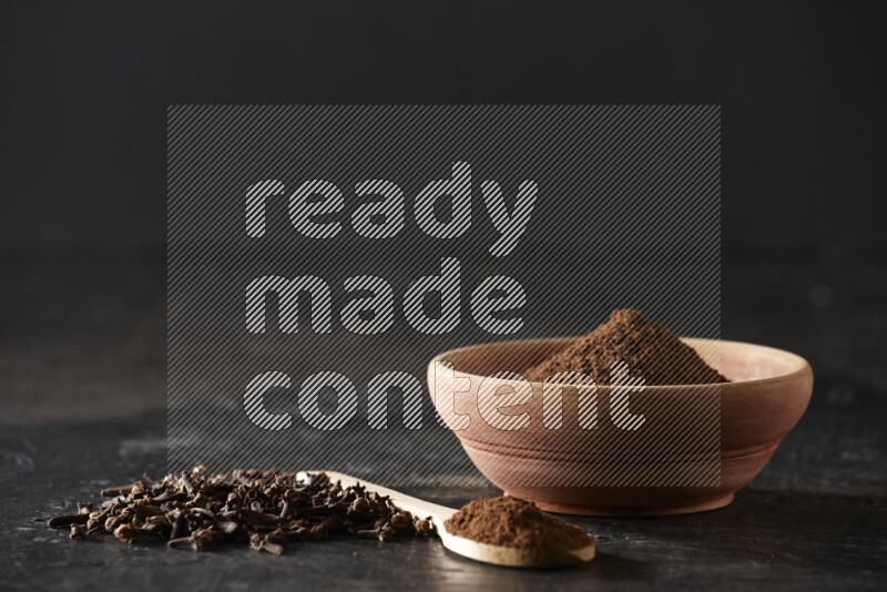 A wooden bowl and a wooden spoon full of cloves powder with spreaded cloves on a textured black flooring