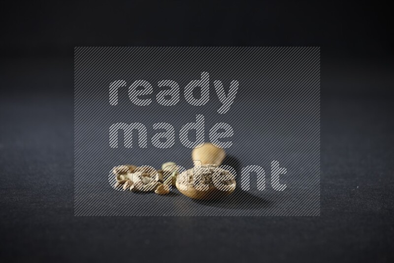 A wooden spoon full of cardamom powder and cardamom seeds beside it on black flooring
