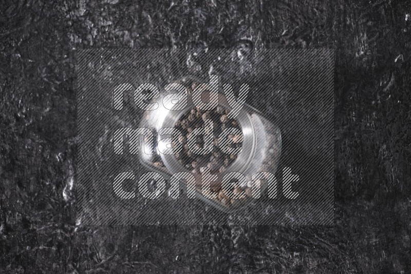 A glass spice jar full of black pepper beads on textured black flooring