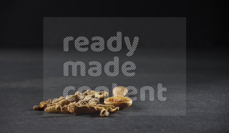 A wooden spoon full of turmeric powder with dried turmeric fingers on black flooring