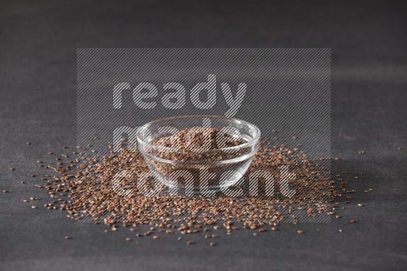 A glass bowl full of flaxseeds surrounded by the seeds on a black flooring