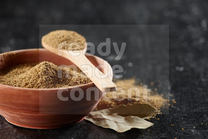 A wooden bowl and spoon full of cumin powder on a textured black flooring