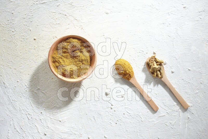 A wooden bowl full of turmeric powder and 2 wooden spoons full of dried turmeric whole finger and turmeric powder on textured white flooring