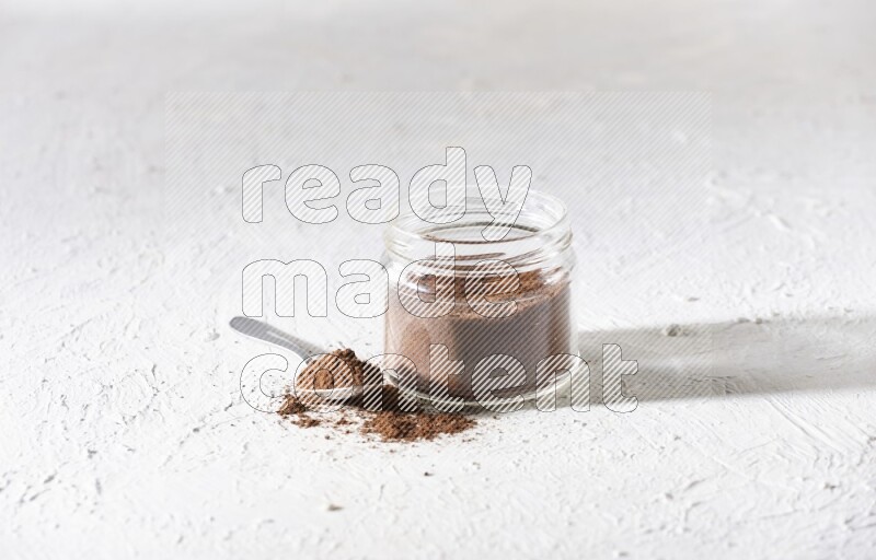 A glass jar full of cloves powder with a metal spoon on a textured white flooring