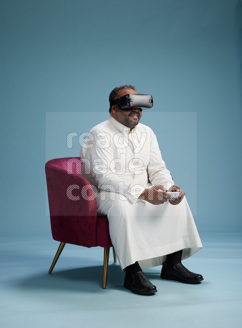 Saudi Man without shimag sitting on chair  gaming with VR and joystick on blue background