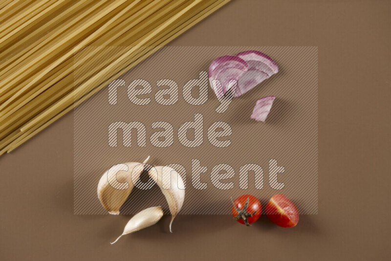 Raw pasta with different ingredients such as cherry tomatoes, garlic, onions, red chilis, black pepper, white pepper, bay laurel leaves, rosemary and cardamom on beige background