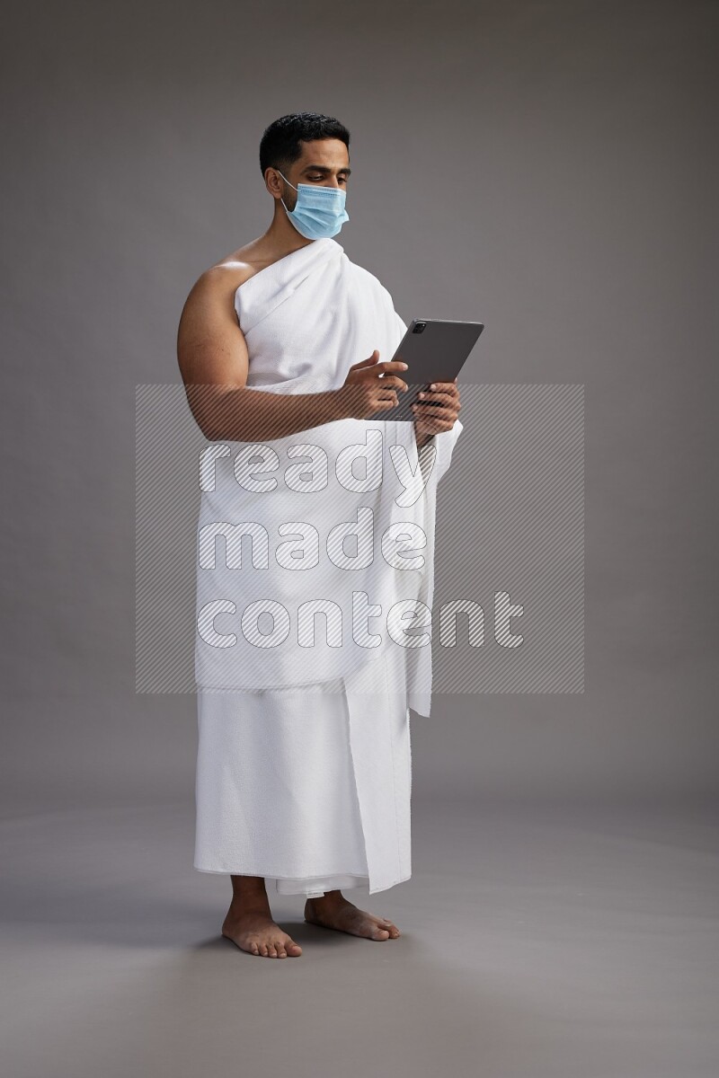 A man wearing Ehram with face mask Standing holding a tablet on gray background